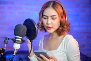 A young smiling female singer wearing headphones with a microphone while recording song in a music studio with colorful lights.