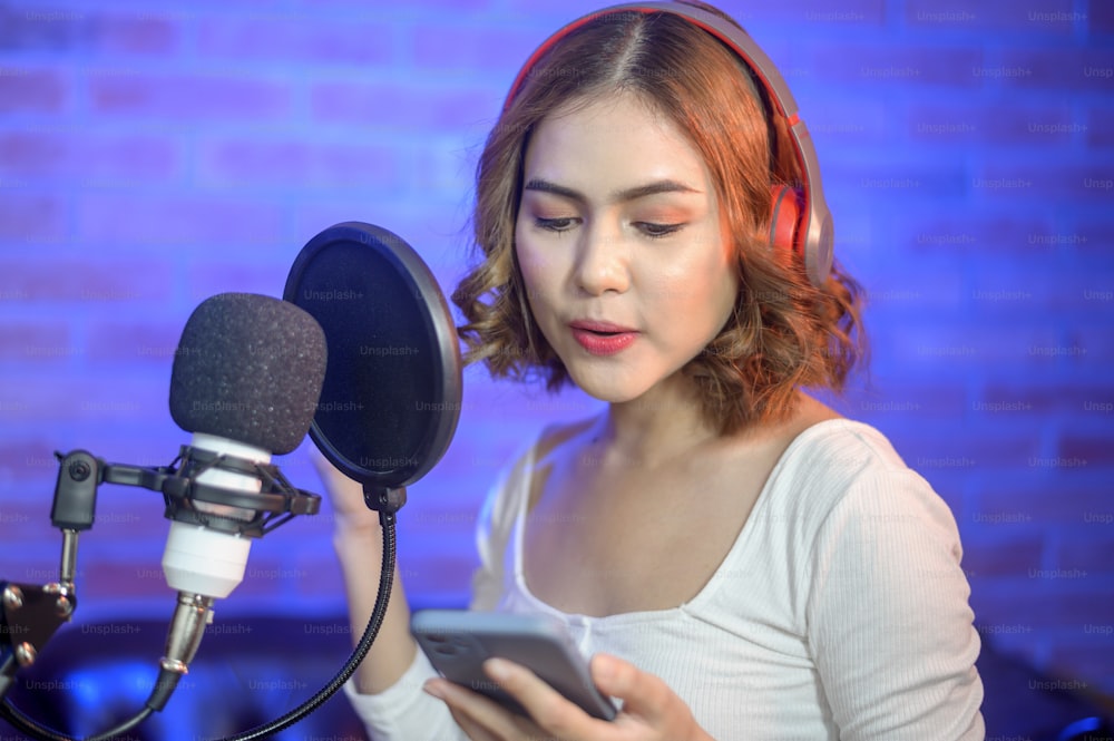 A young smiling female singer wearing headphones with a microphone while recording song in a music studio with colorful lights.