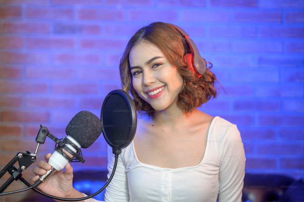 A young smiling female singer wearing headphones with a microphone while recording song in a music studio with colorful lights.