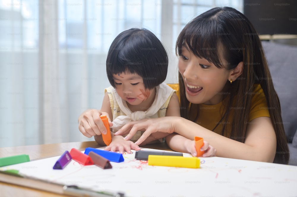 A young mom helping daughter drawing with colored pencils in living room at home.