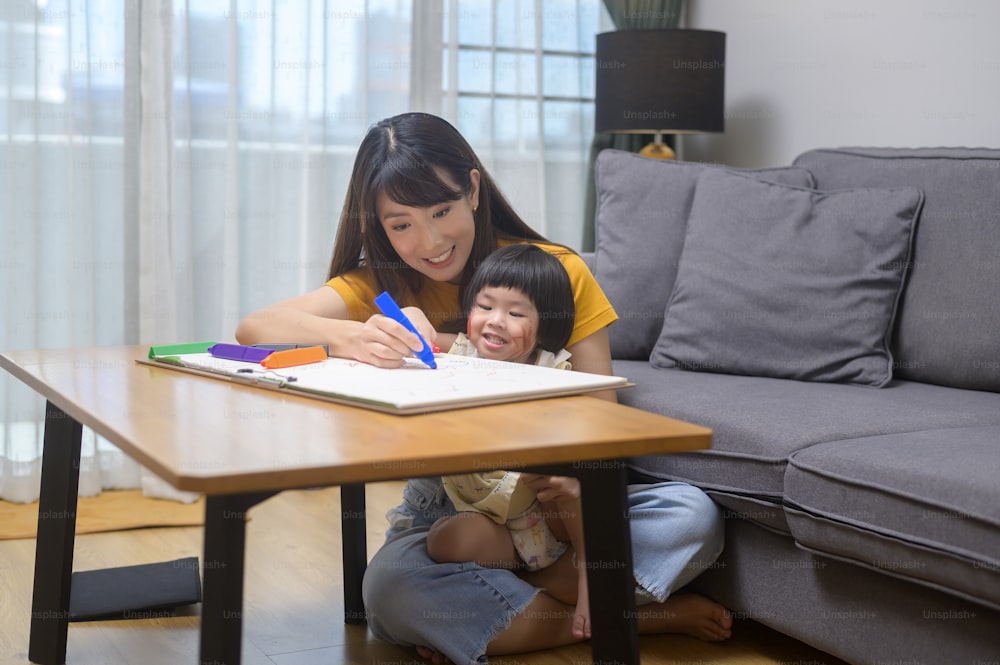 Eine junge Mutter hilft ihrer Tochter beim Zeichnen mit Buntstiften im Wohnzimmer zu Hause.