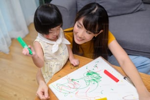 Une jeune maman aidant sa fille à dessiner avec des crayons de couleur dans le salon à la maison.