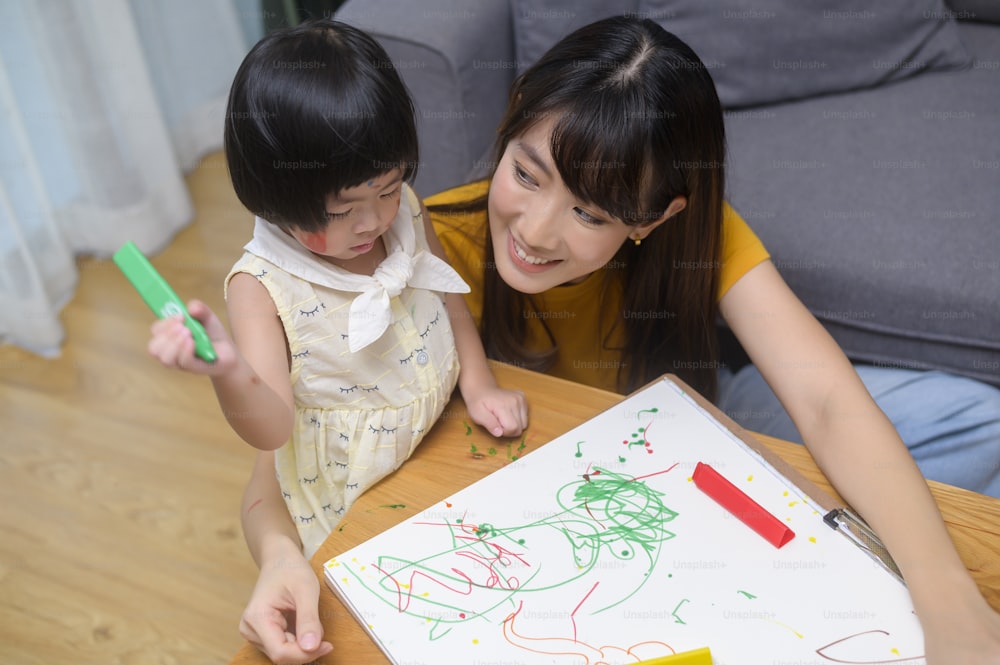 딸이 집 거실에서 색연필로 그림을 그리는 것을 돕는 젊은 엄마.