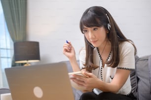 Uma bela jovem mulher usando fone de ouvido está fazendo chamada de videoconferência via computador em casa