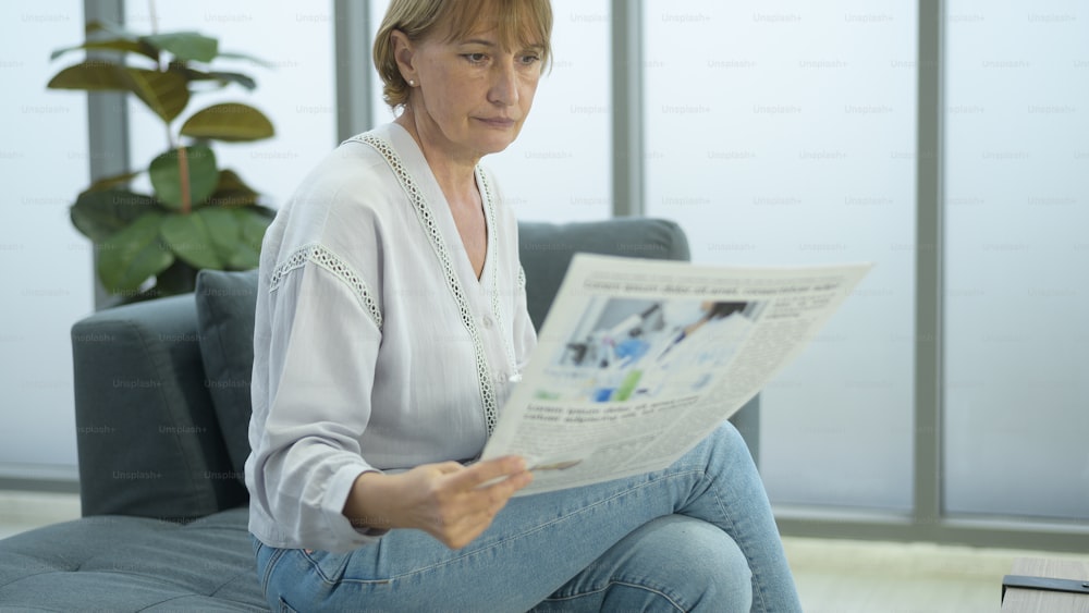 Mulher idosa caucasiana feliz está relaxando, lendo jornal na sala de estar