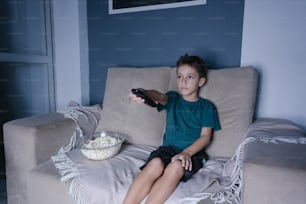 Menino pequeno no sofá assistindo TV e comendo pipoca à noite na sala de estar