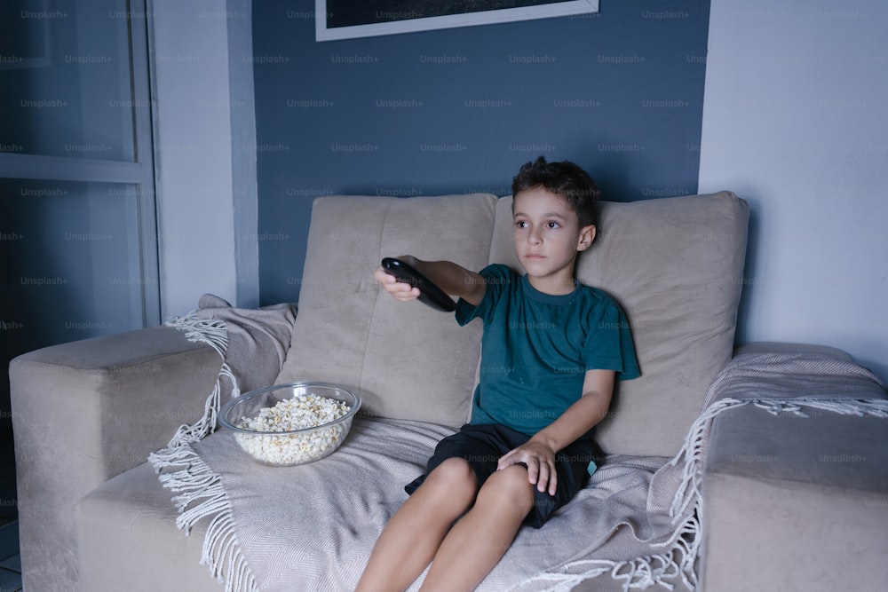 Kleiner Junge auf der Couch, der nachts im Wohnzimmer fernsieht und Popcorn isst