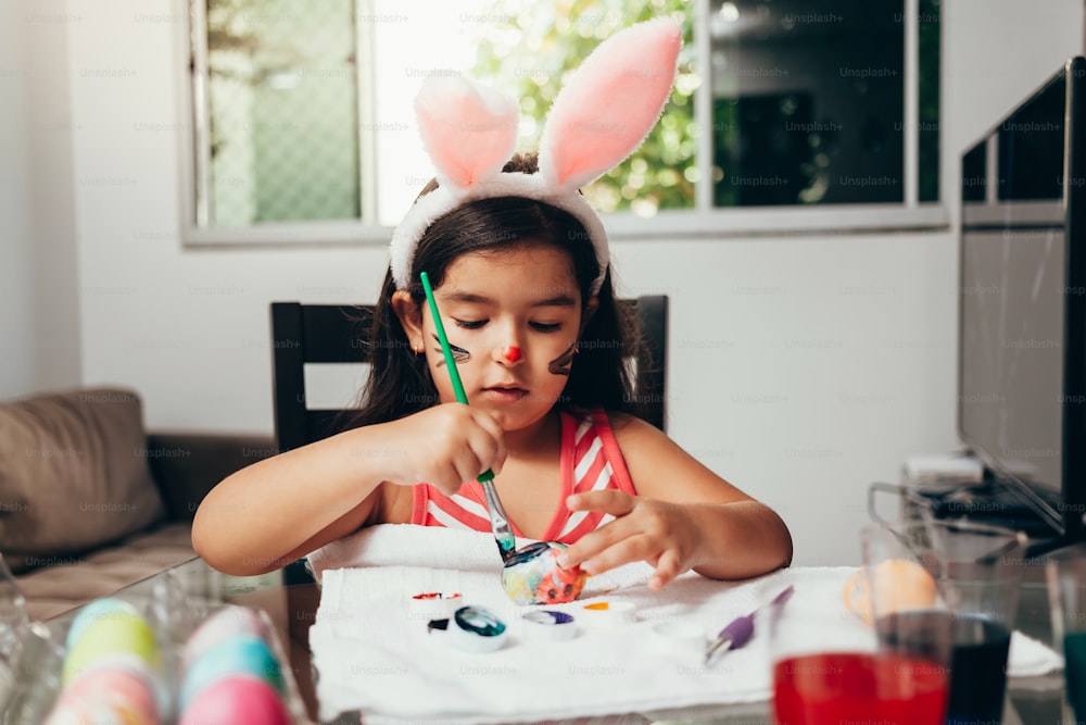 Frohe Ostern! Ein wunderschönes Kind, das Ostereier bemalt. Glückliche Familie, die sich auf Ostern vorbereitet. Süßes kleines Kind, das am Ostertag Hasenohren trägt