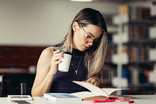 Young woman studying at table in library. Holding a cup of coffee