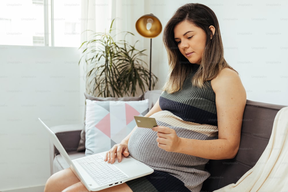 Concepto de compra en línea. Mujer embarazada con computadora portátil y tarjeta de crédito comprando artículos para bebés en Internet