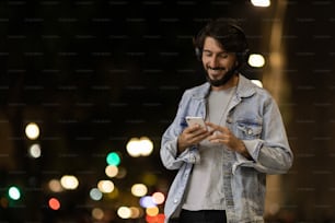 Front view of young man using a smartphone and headphones at night time with city view landscape in the background. High quality photo Mobile phone, technology, concept. High quality photo