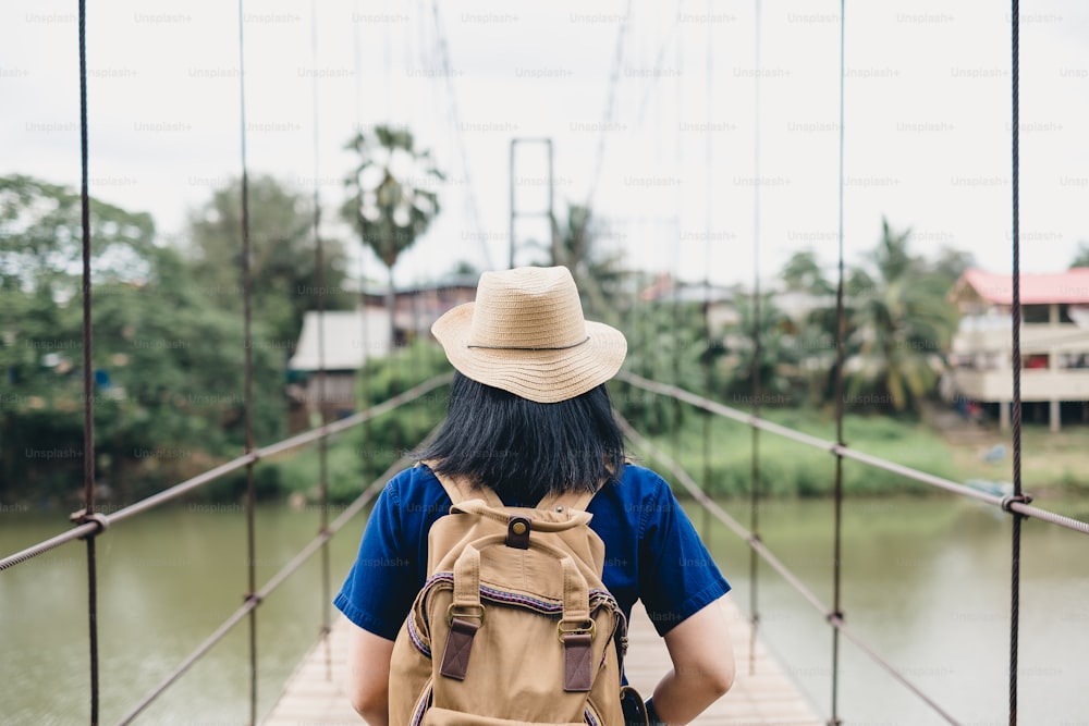 Donna asiatica viaggiatrice con zaino a ponte pedonale attraverso il fiume in campagna, vacanza estiva in viaggio