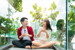 魅力的なアジアのカップルのカジュアルなドレスは、陽気な笑いと笑顔の手遊びを楽しみ、現代の庭で一緒にスマートフォンでオンラインモバイルゲームを競います