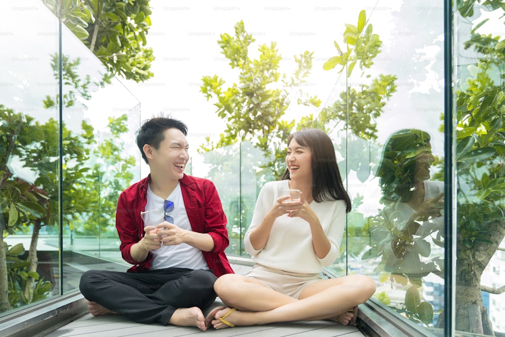 Atractiva pareja asiática vestido casual disfrutar de la risa alegre y la sonrisa juego de mano en línea juego móvil juntos competencia en teléfono inteligente juntos en el jardín moderno