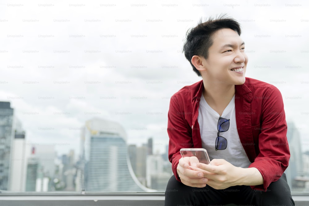 若いアジアの男性カジュアルリラックス赤シャツ手持ちガラススマートフォン都市ダウンタウンのビジネステクノロジーのアイデアコンセプトの背景