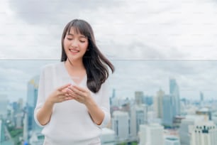 portrait of attractive asian long black hair casual dress tootly smile hand use smartphone with background of cityscape and white light sky copyspace freedom and cheerful emotional