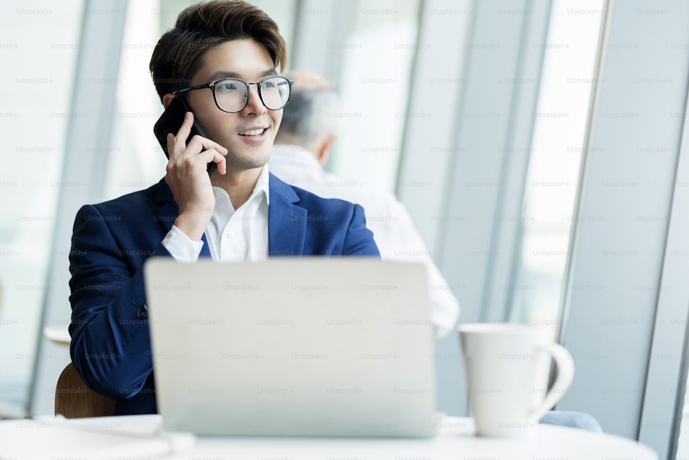 agência criativa óculos inteligentes asiáticos masculino conversa formal de pano com smartphone freelance trabalhando com laptop na área de coworking espaço de escritório com frescor com fundo de escritório desfocado