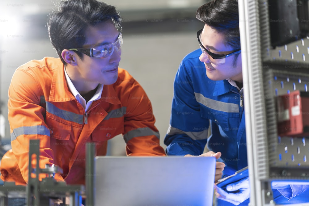 工場で機械のそばに立って議論をしているアジアの男性エンジニアの専門家、2人のアジア人の同僚が説明し、機械のプロセスcurcuitマザーボードを解決します
