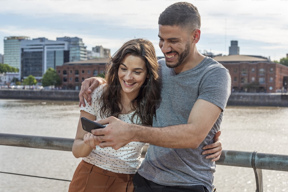 Happy young couple holding a modern smartphone in hands. They are by the river in a beautiful city.