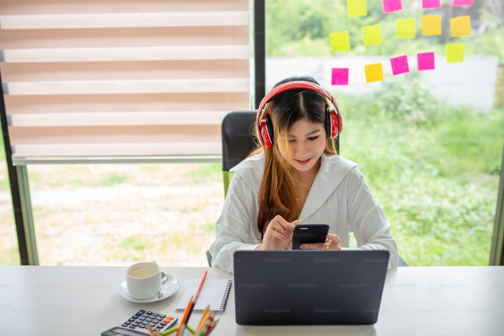 Portrait of young asian woman listening to music with headphones and using laptop computer and mobile phone at office.