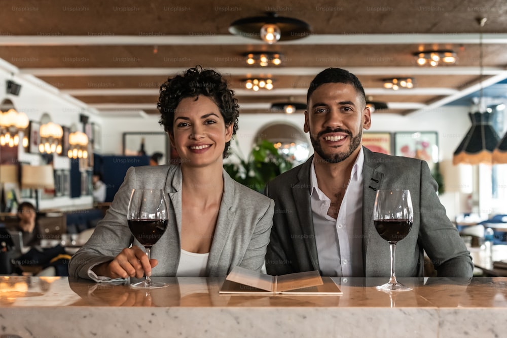 Young lovely couple enjoying at the bar in the evening. They are sitting at the bar with a glass of wine looking at the camera.