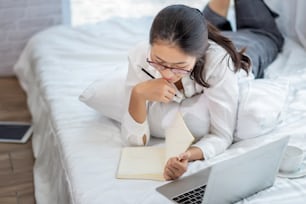 Young asian woman working on laptop and take notes on bed at home.