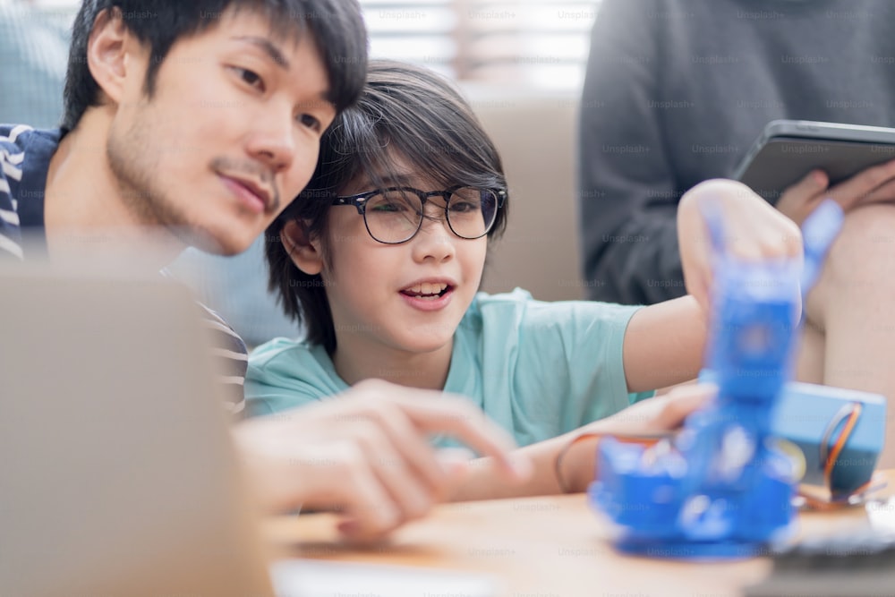 Concept d’étude à la maison, enfant garçon asiatique avec le bras de robot de contrôle réussi du père et de la mère sur la tablette numérique et le clavier d’ordinateur, famille heureuse recevant une leçon de robotique de contrôle de codage à la maison