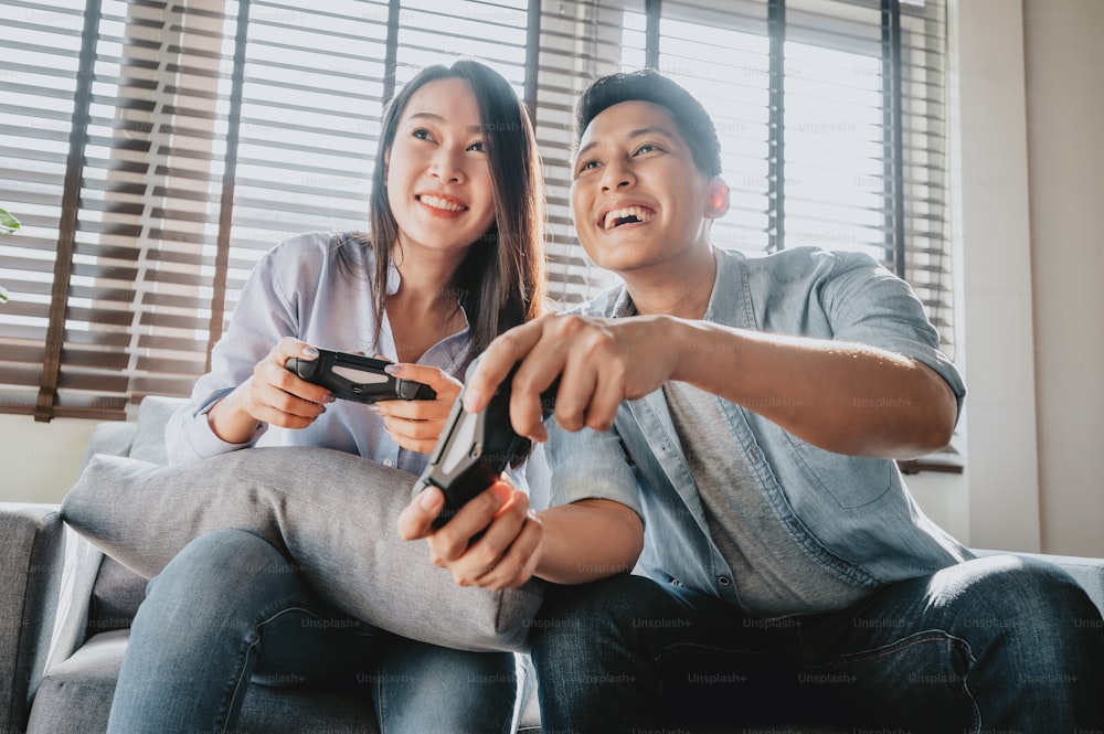 Happy Asian couple using joystick controller playing video games together while sitting on sofa  in their living room and having fun