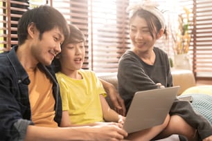 happiness asian family sunday morning mom dad son sitting together using laptop play game online together with exited fun cheerful casual relax relation in living room home interior background