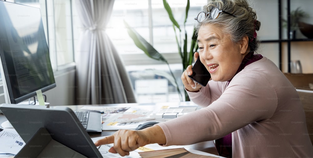 Elderly senior asian female freelancer in casual clothes typing on laptop keyboard while talking on smartphone sitting at desk and busy working in home office