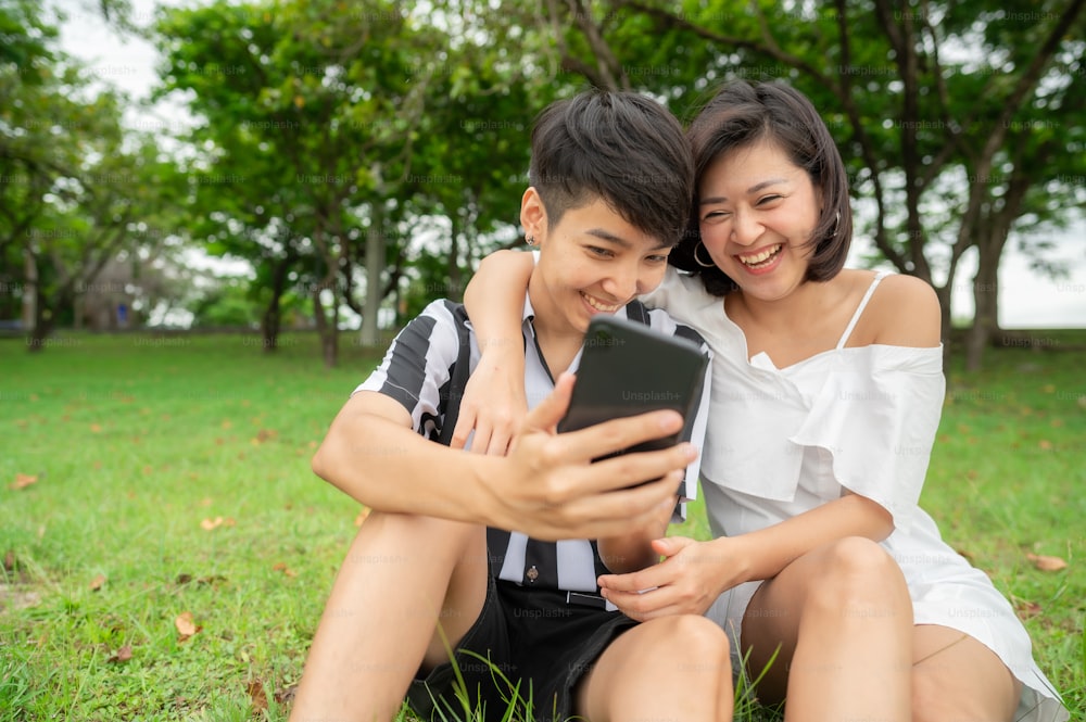 Happy Laughing Asian Lesbian Couple Sitting Outdoor In The Park While Using Mobile Phone Photo
