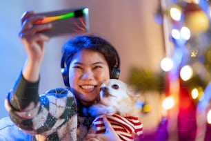 若いアジアの女性の大人の手は、自宅でクリスマスと新年のお祝いの週末の休日の休暇に笑顔と幸福感を持つ彼女の小さな子犬の犬と自撮り写真を撮るスマートフォンを使用しています