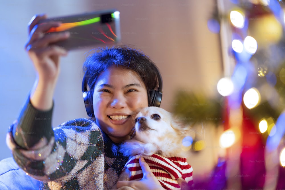 젊은 아시아 여성 성인 손은 집에서 크리스마스와 새해 축제 주말 휴가 휴가에서 미소와 행복감을 가진 그녀의 작은 강아지와 셀카 사진을 찍는 스마트폰을 사용합니다.