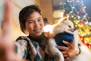 젊은 아시아 여성 성인 손은 집에서 크리스마스와 새해 축제 주말 휴가 휴가에서 미소와 행복감을 가진 그녀의 작은 강아지와 셀카 사진을 찍는 스마트폰을 사용합니다.