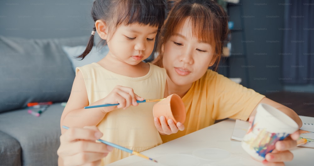 Une maman de la famille Happy Asia enseigne au pot en céramique de peinture pour les tout-petits filles en s’amusant sur la table, dans le salon de la maison. Passer du temps ensemble, Distanciation sociale, Quarantaine pour la prévention du coronavirus.