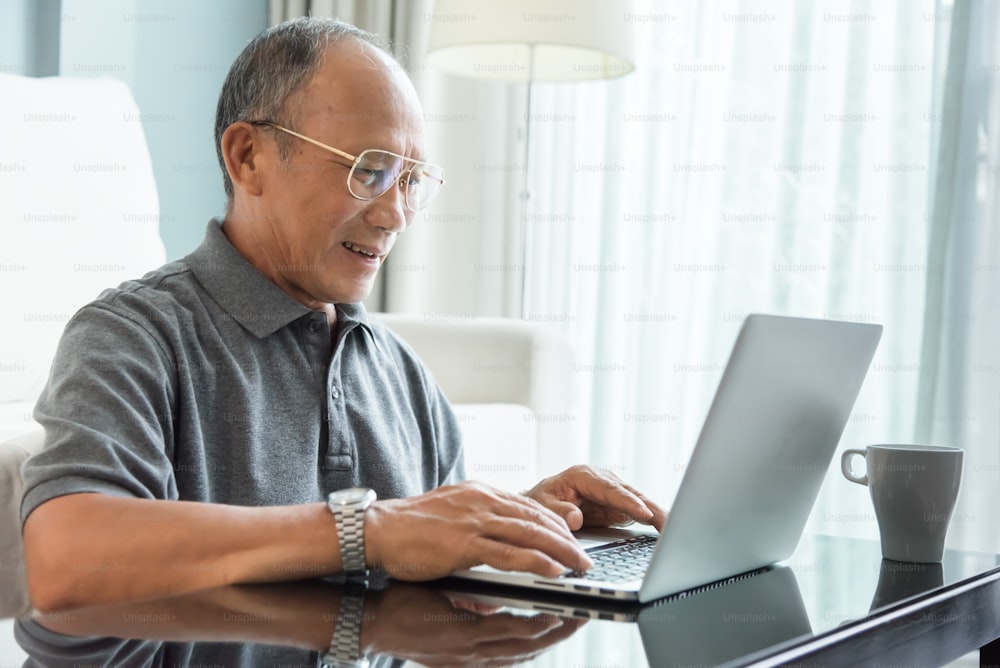 그의 집에서 노트북을 사용하는 편안한 아시아 노인. 행복한 노인 남성이 웃고 있다. 퇴직