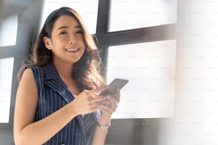Attractive Modern Asian businesswoman using mobile phone with smiling and standing near window in office. Confident Small business female entrepreneur looking at camera while holding smartphone.