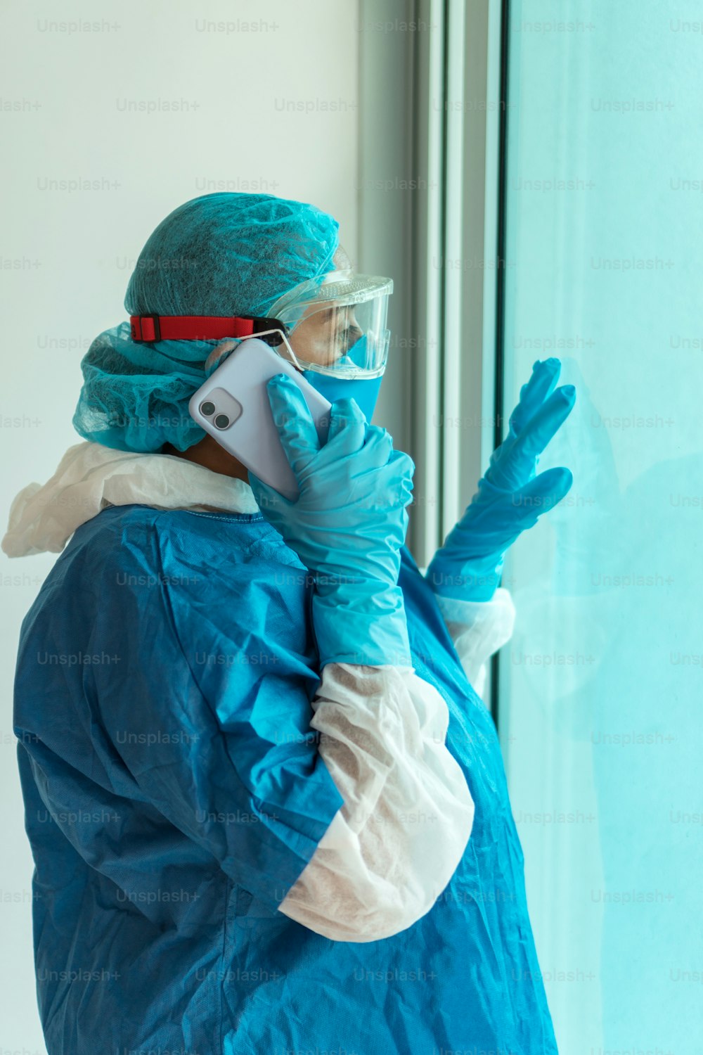 Side view of a woman in a protective gown with a mask and goggles talking on her cell phone and looking out her window while working in the hospital during the coronavirus outbreak