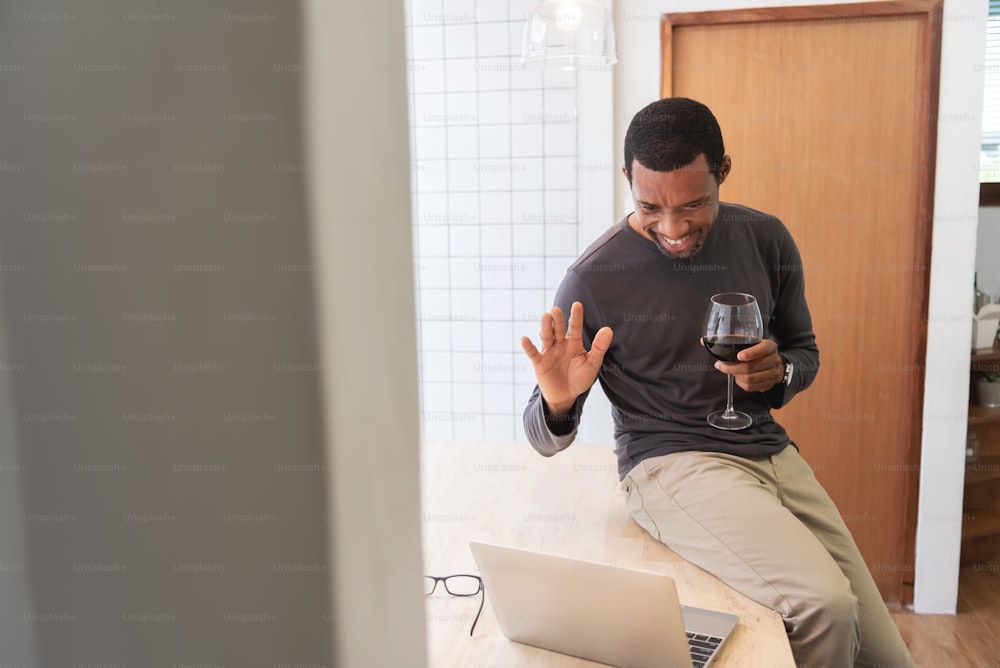 Smiling African American man enjoying and waving hand with his friends on notebook. Happy Black male doing online celebration with toasting red wine via video call on laptop during quarantine at home