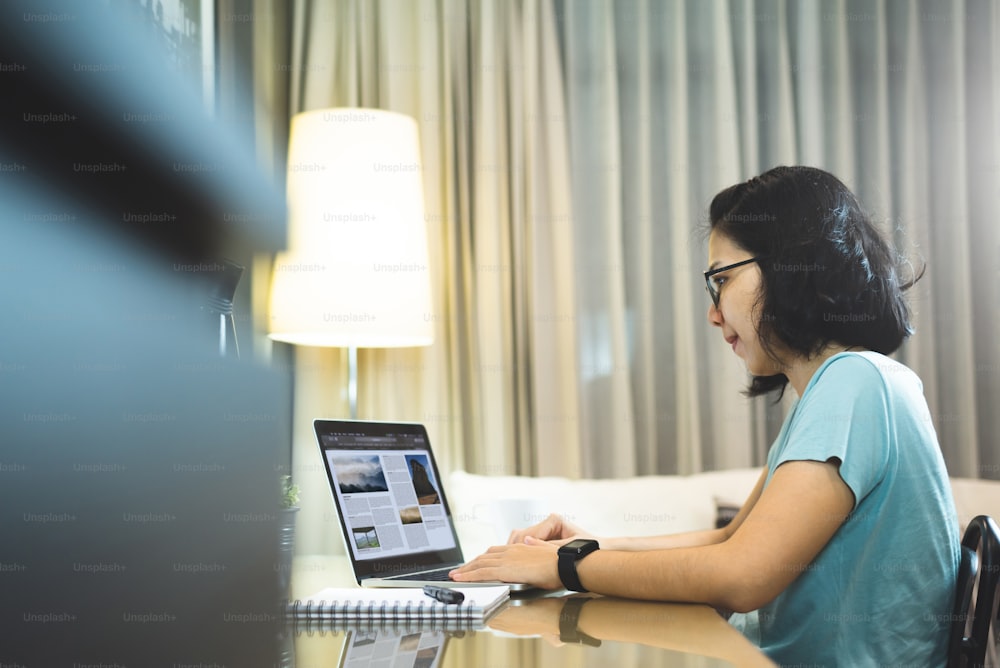 Asian woman Freelancer Designer wear eyeglasses working on laptop computer in night time. Young Female student studying and doing online learning at home. Social distancing, New normal life concept.