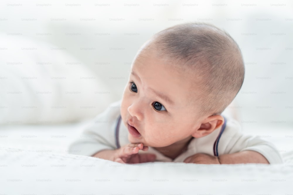 Unschuldige junge süße asiatische Neugeborene lernen, auf dem Bett im Schlafzimmer zu krabbeln. Das Baby lächelt und schaut glücklich und freudig nach rechts. Säuglingsgesundheits- und Babyentwicklungskonzept.