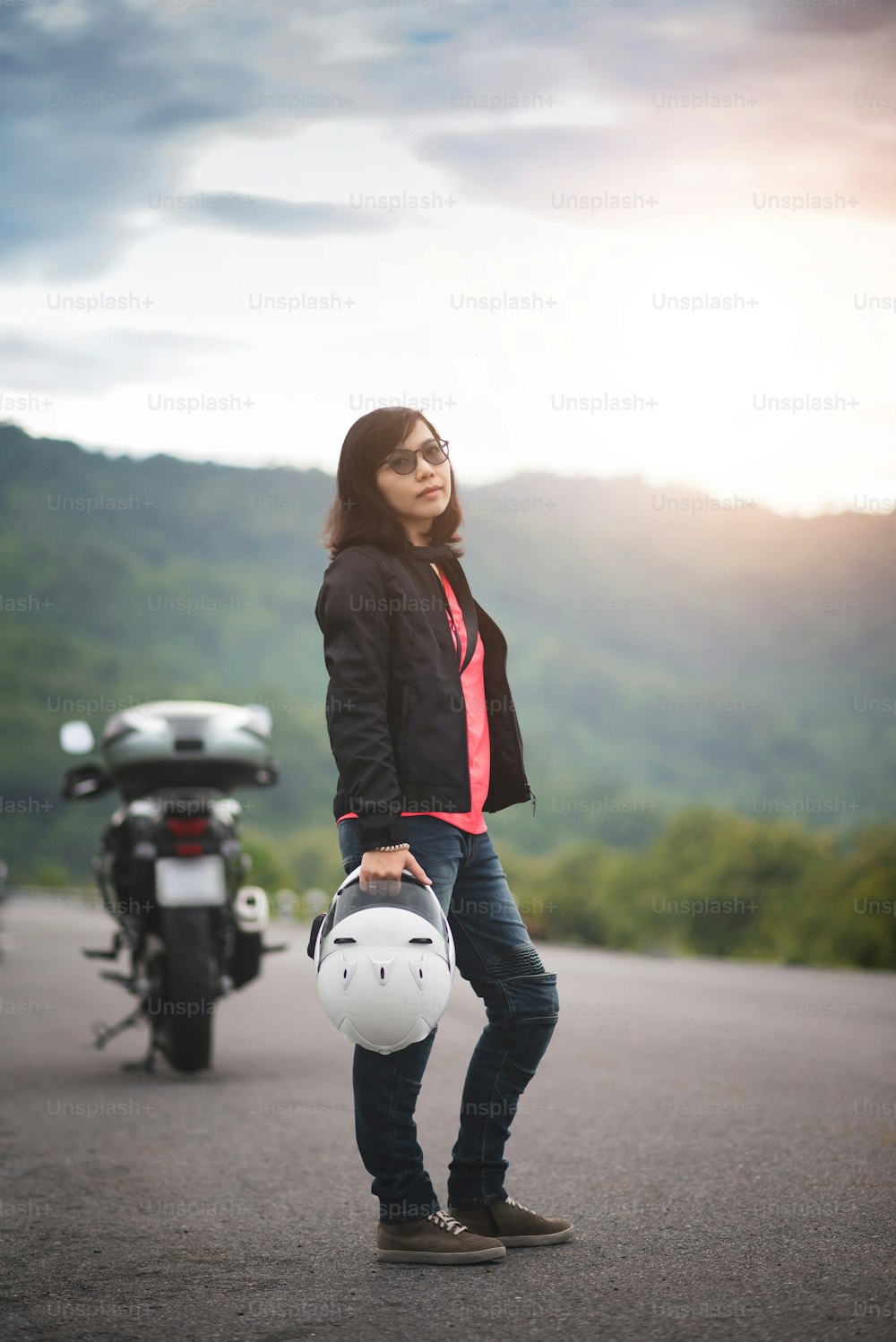 Asian woman wear sunglasses and protective jacket is holding a white helmet against a motorcycle and mountain background. Biker, Rider, Travel.