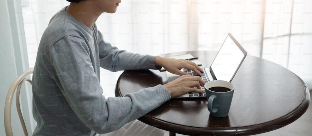 Asiatisch Junger Mann im Pullover benutzt einen Laptop auf einem runden Holzschreibtisch an seinem Arbeitsplatz in seinem Haus. Tippen, Jobsuche, Verbindung mit drahtloser Technologie.