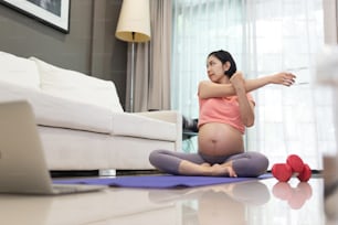 Attractive Asian Pregnant woman in pink sport clothing stretching arms at home. Young Female doing yoga on exercise mat while her pregnancy in  living room. Healthy Single mom work out in online class