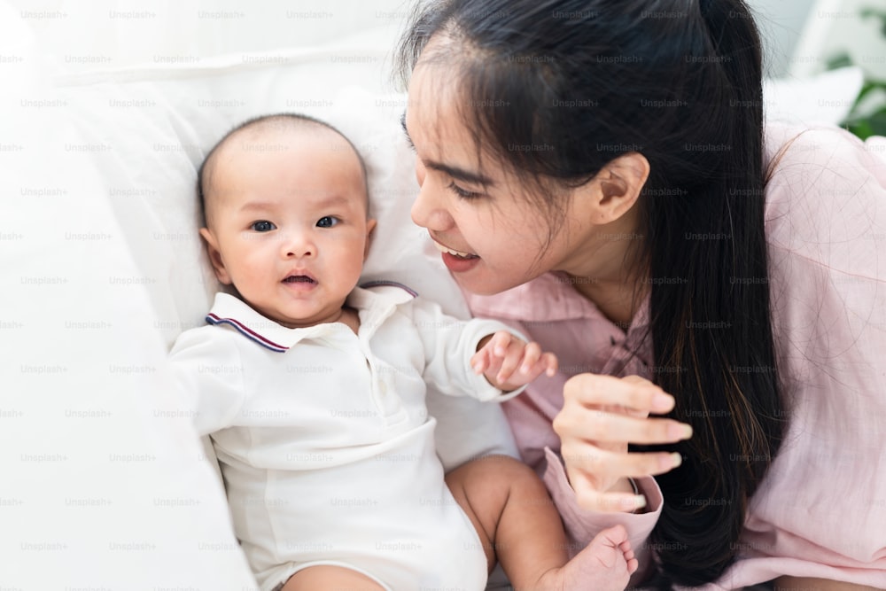 Asian cute baby sit on bed in warm house. Mother looks and smiles to happy little boy. Beautiful lovely infant newborn feel safe to be with family. Warm touch happiness and healthy family concept.