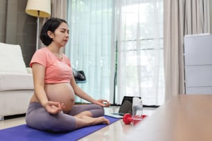 リビングルームで蓮華の姿勢でヨガのエクササイズをしている魅力的な妊婦の肖像画。ピンクのスポーツウェアを着た健康な若いアジアの母親と目を閉じて自宅でマインドフルネス瞑想。