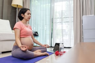 Porträt einer attraktiven schwangeren Frau, die Yoga-Übungen im Lotussitz im Wohnzimmer macht. Gesunde junge asiatische Mutter in rosa Sportbekleidung mit geschlossenen Augen Achtsamkeitsmeditation zu Hause.