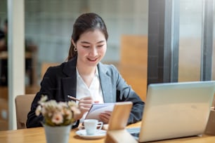 Jovem empresária asiática bonita trabalhando em laptop e tomando notas enquanto sentada à mesa no escritório.