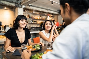 Gruppo di giovani migliori amici interrazziali persone sedute al ristorante al coperto che mangiano cibo insieme a pranzo. Donne che parlano con l'uomo asiatico del pasto con sorriso. Si sentono felici e si godono il pasto.