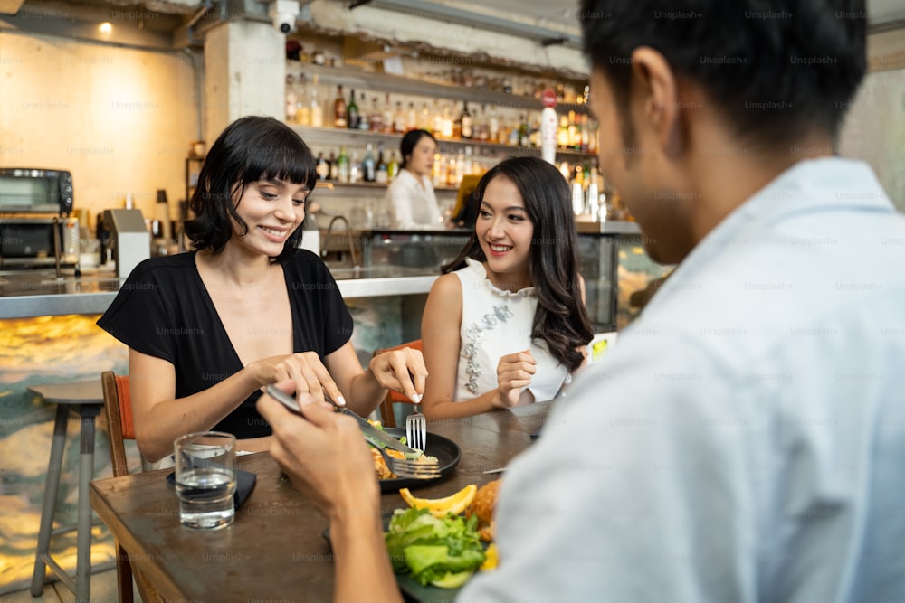 Group of young interracial best friend people sitting indoor restaurant eating food together for lunch. Women talking to Asian man about meal with smile. They are feeling happy and enjoying the meal.